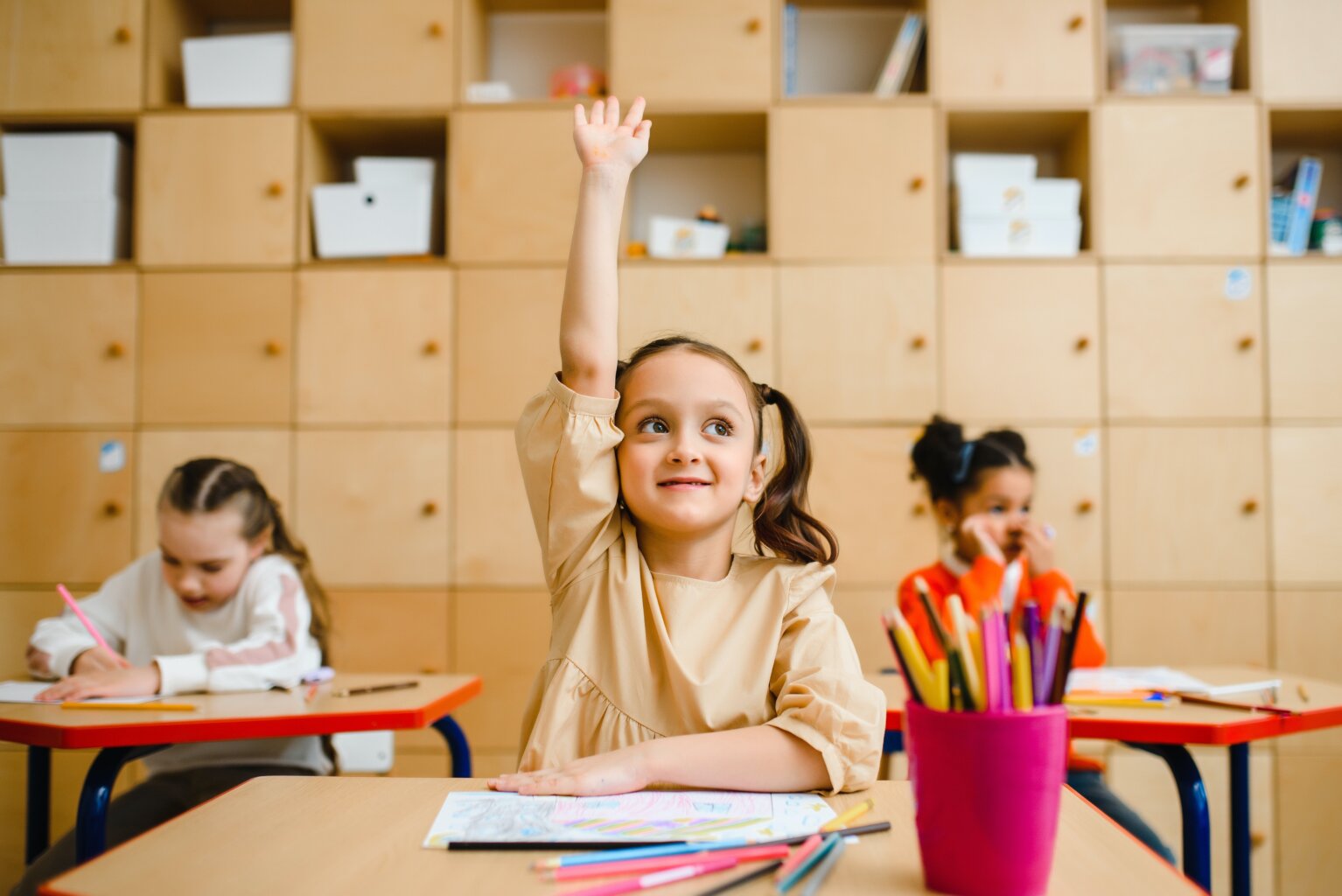 A young girl raising her hand in class with her school supplies spread out around her.