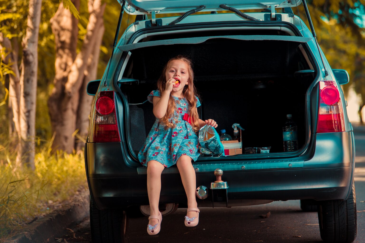 A girl sitting in the trunk of a car while on a road trip and eating snacks.