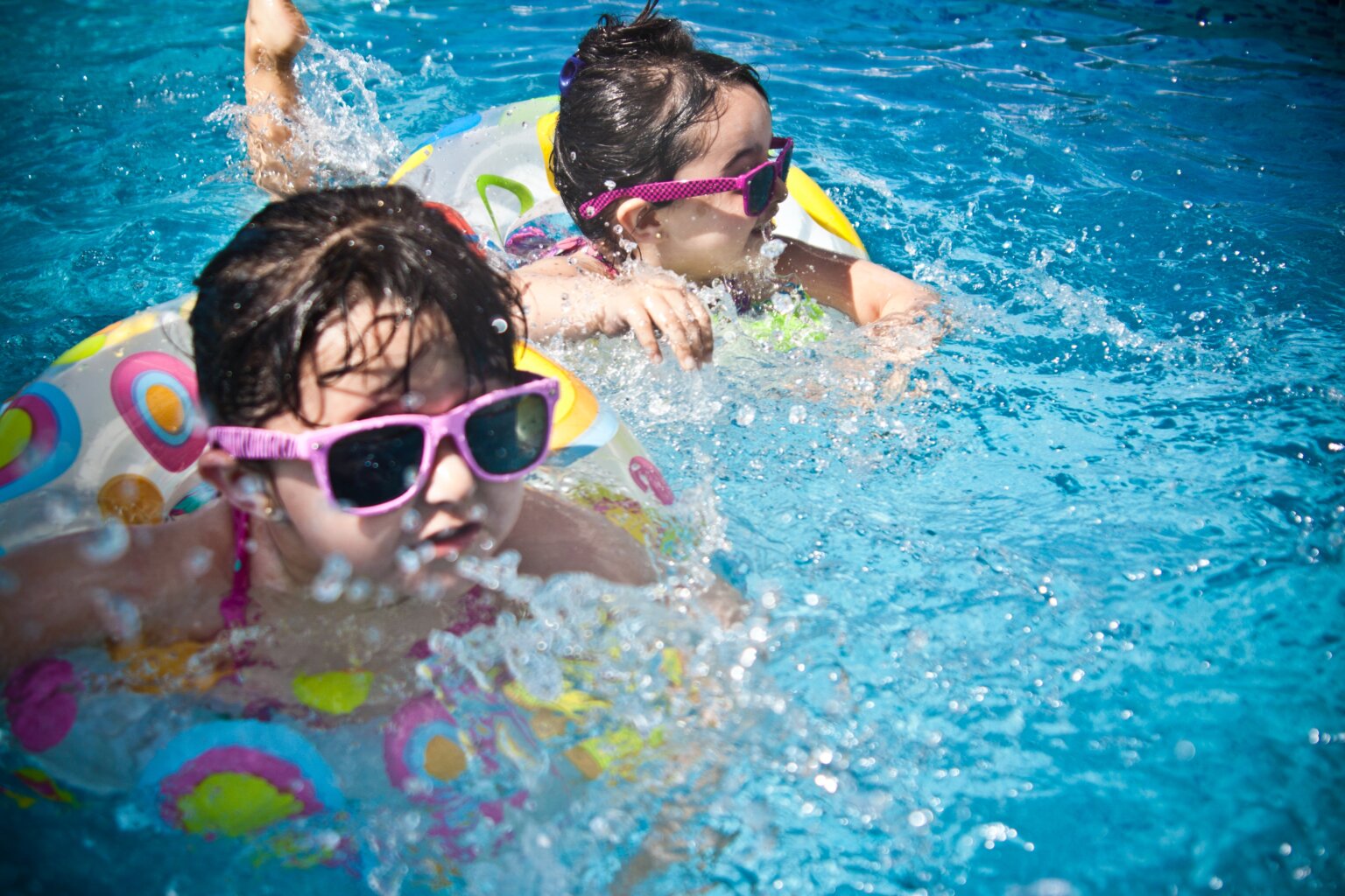 Two girls swimming in a pool with floaties and pink sunglasses.