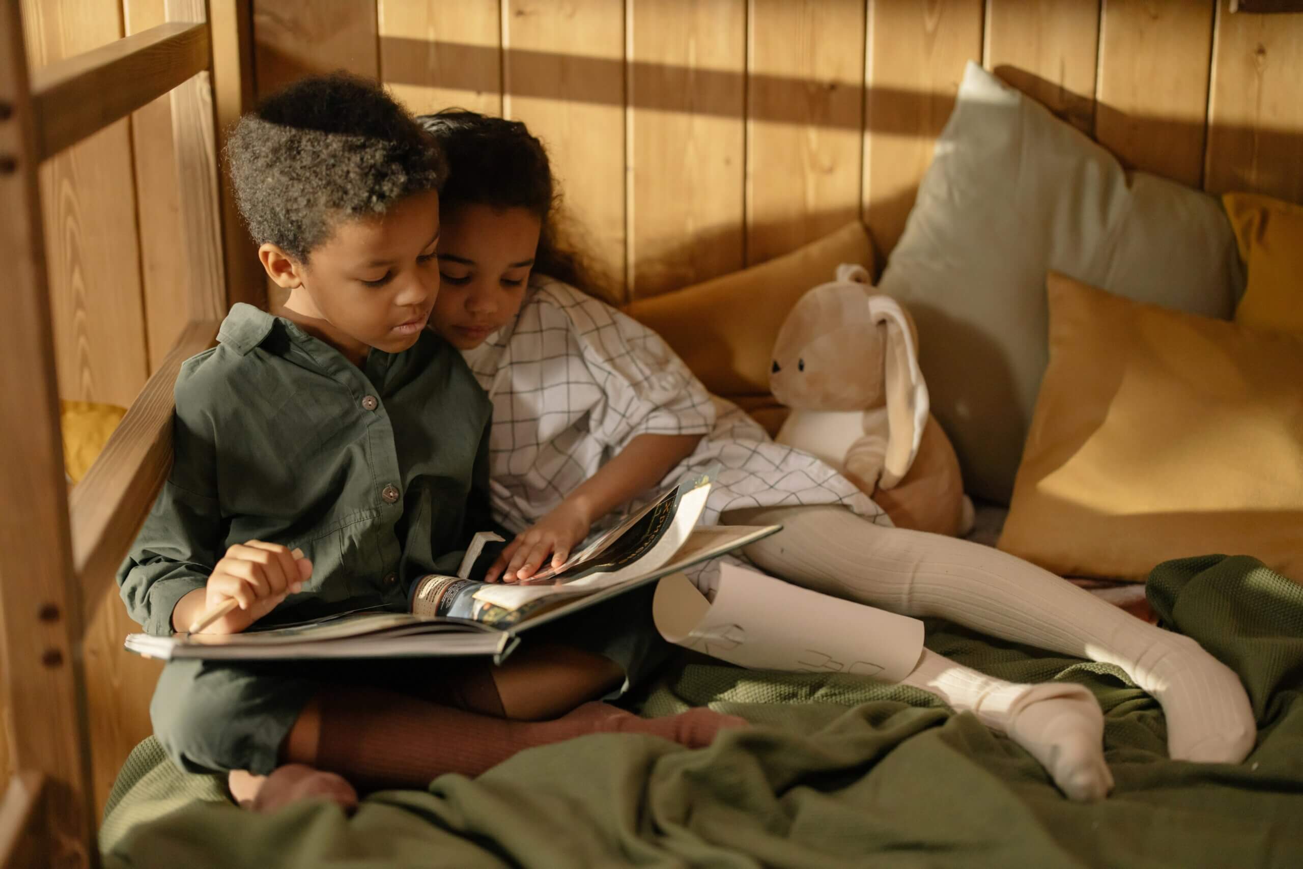 A boy and a girl reading a book on a bed next to a stuffed toy rabbit.