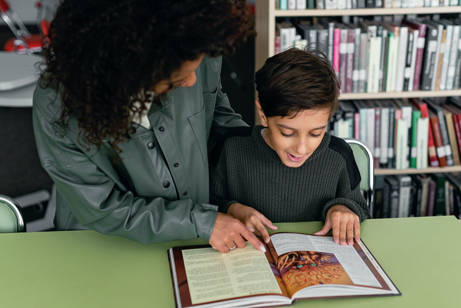 Woman helping a boy read a book in the library.
