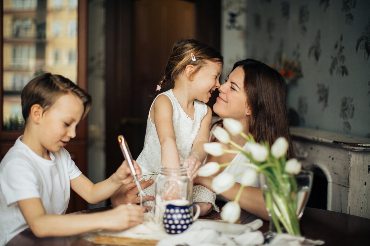 A mom and her daughter touching their noses together while the mom, daughter, and son mix flour at a table.