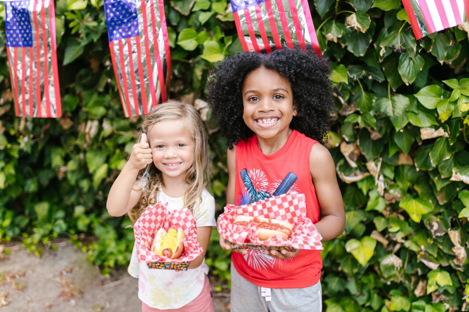 Two girls holding hot dogs while standing in front of American flag party decorations.