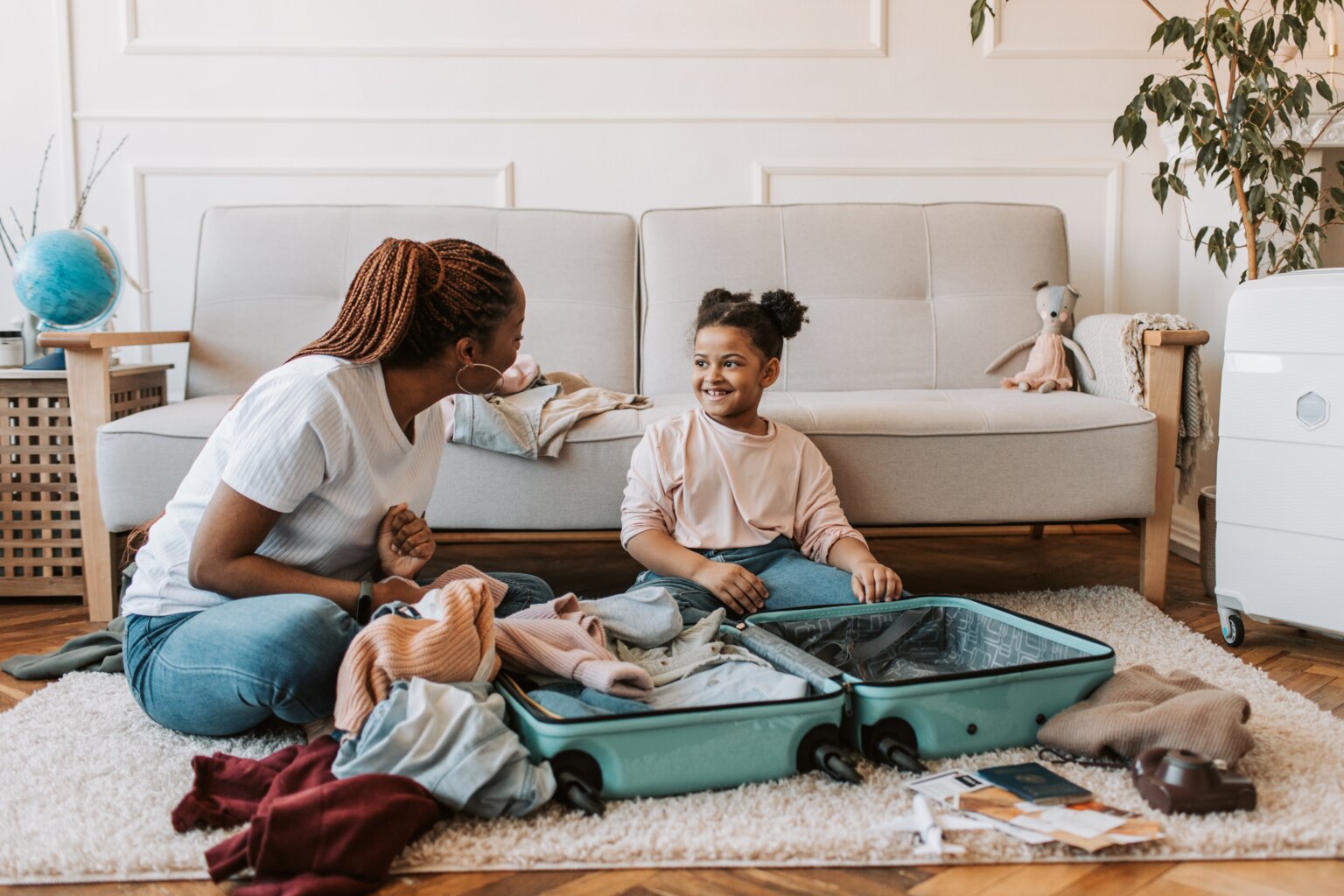 A mom and daughter sitting on the floor of a living room and packing a suitcase.
