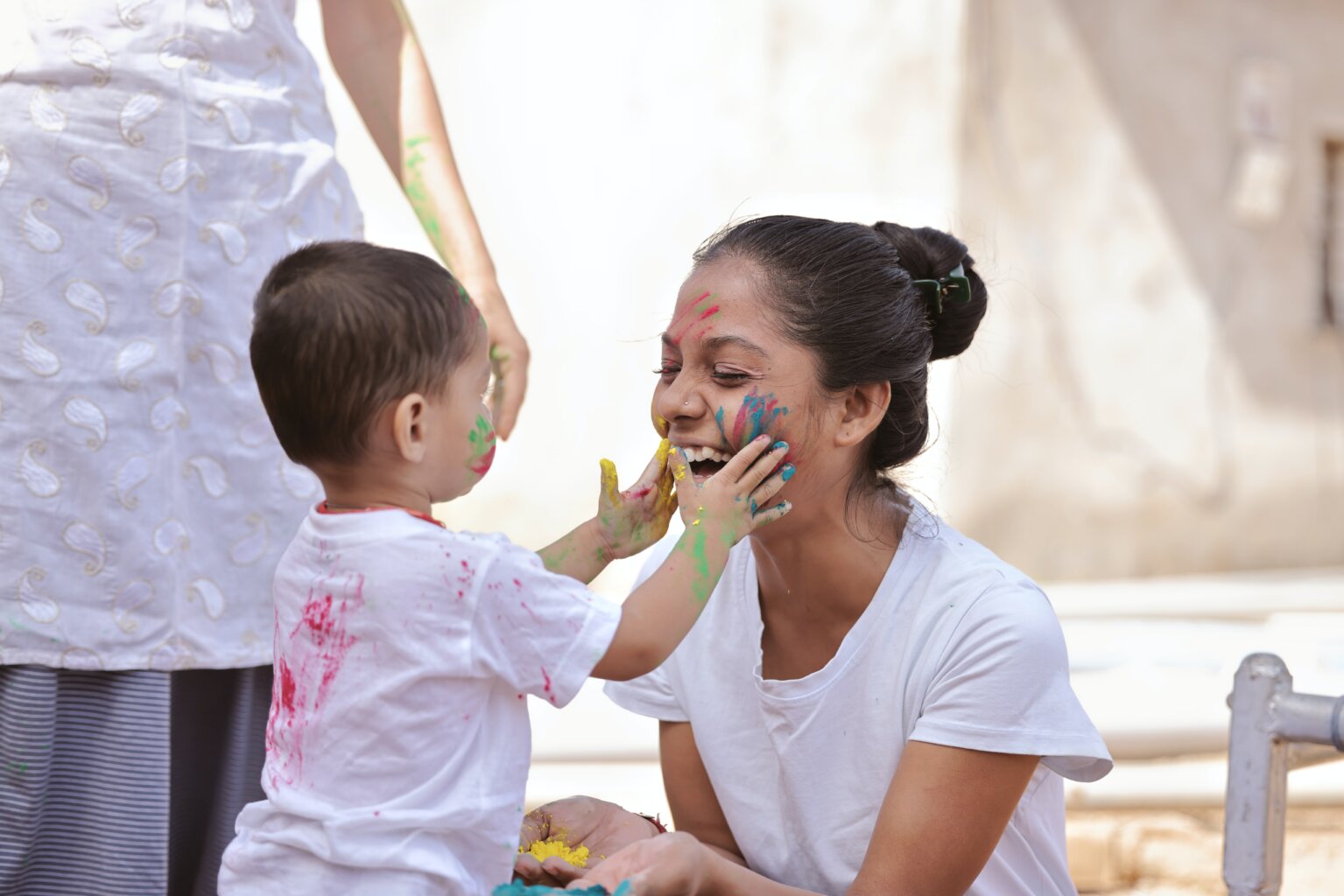 A mom and son who are both covered in paint while the toddler holds his mom’s cheeks.