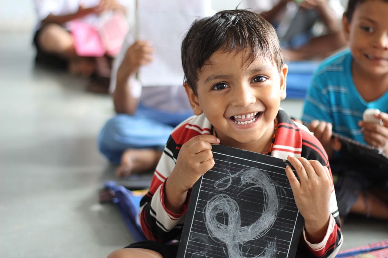 A smiling boy sitting with his classmates at school and holding a chalkboard with scribbles.