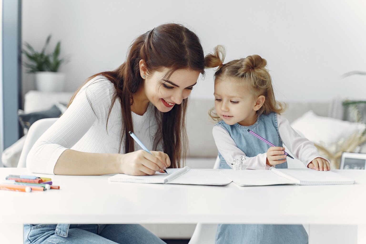 A mom teaching her daughter at a desk while they both write in notebooks.