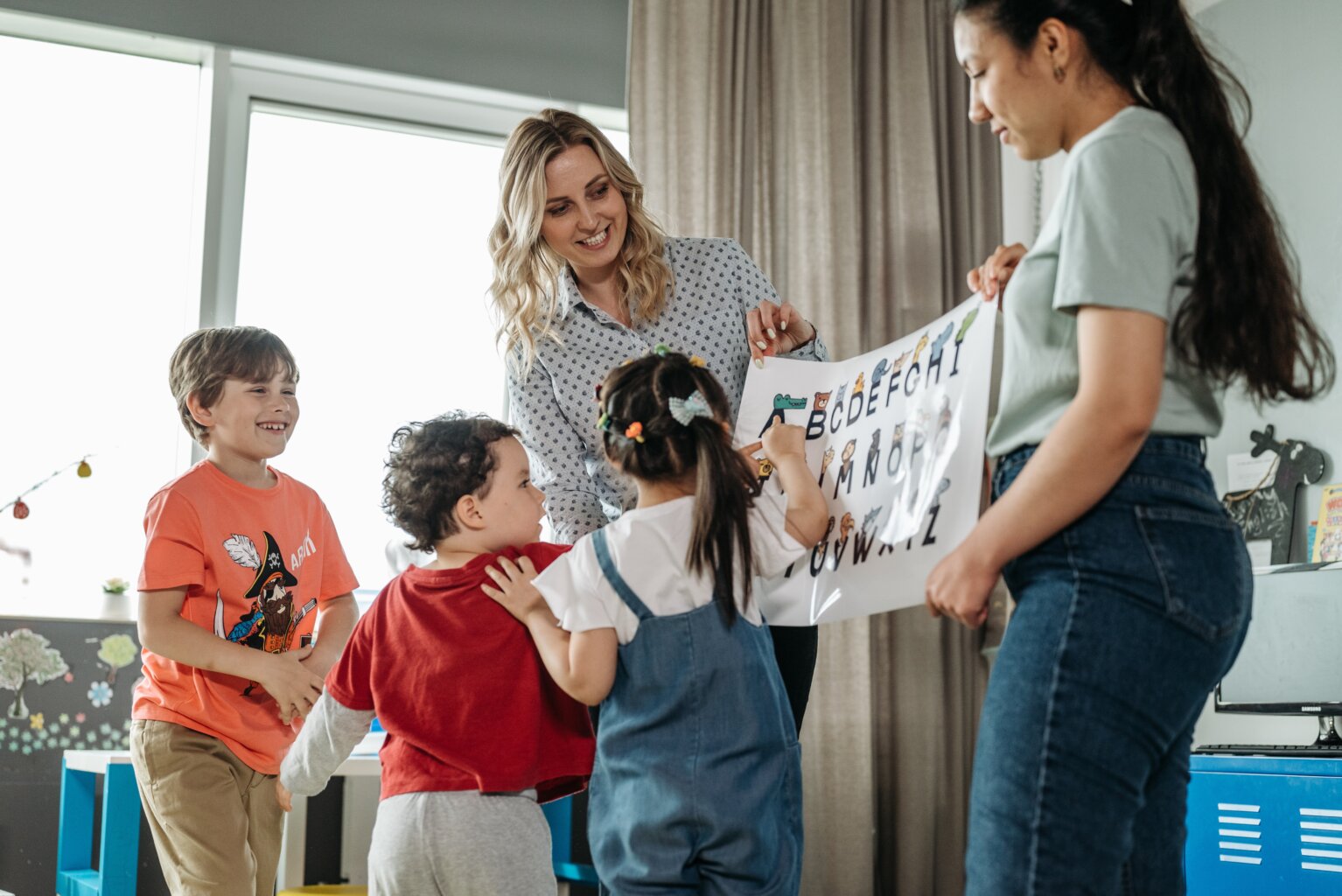 A mom and teacher holding an alphabet poster while the kids point at letters.