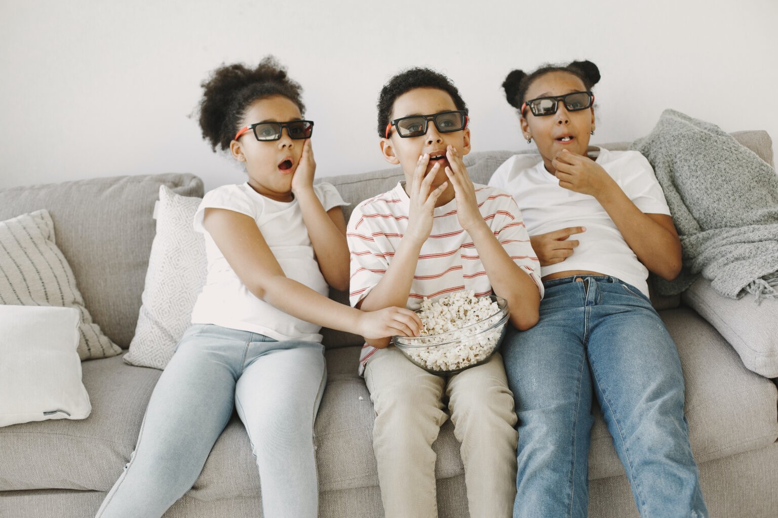 Three kids, two girls and one boy, looking surprised while eating popcorn and watching a movie.