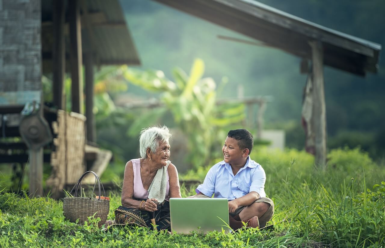 A boy asking his grandmother family history questions in a field with a laptop.
