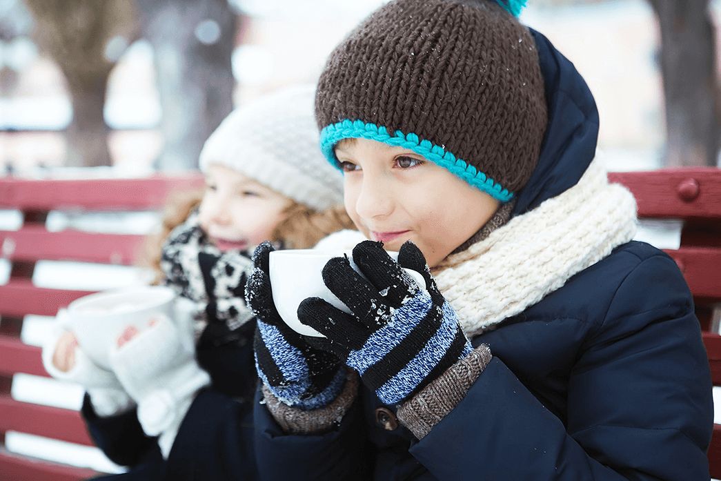 A brother and sister drinking hot chocolate on a park bench in the snow while participating in fun winter activities.