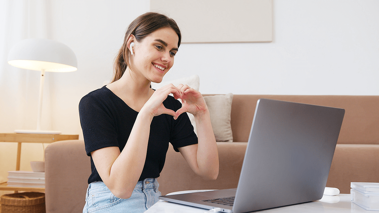A woman signing a heart to a computer camera while on a video call to reconnect with friends.