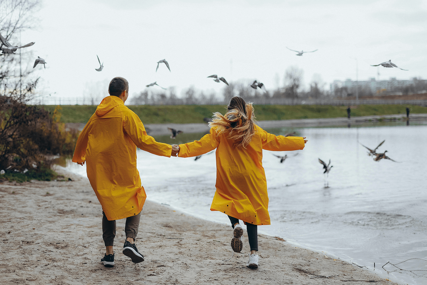 A couple in yellow raincoats running on the shore chasing birds.