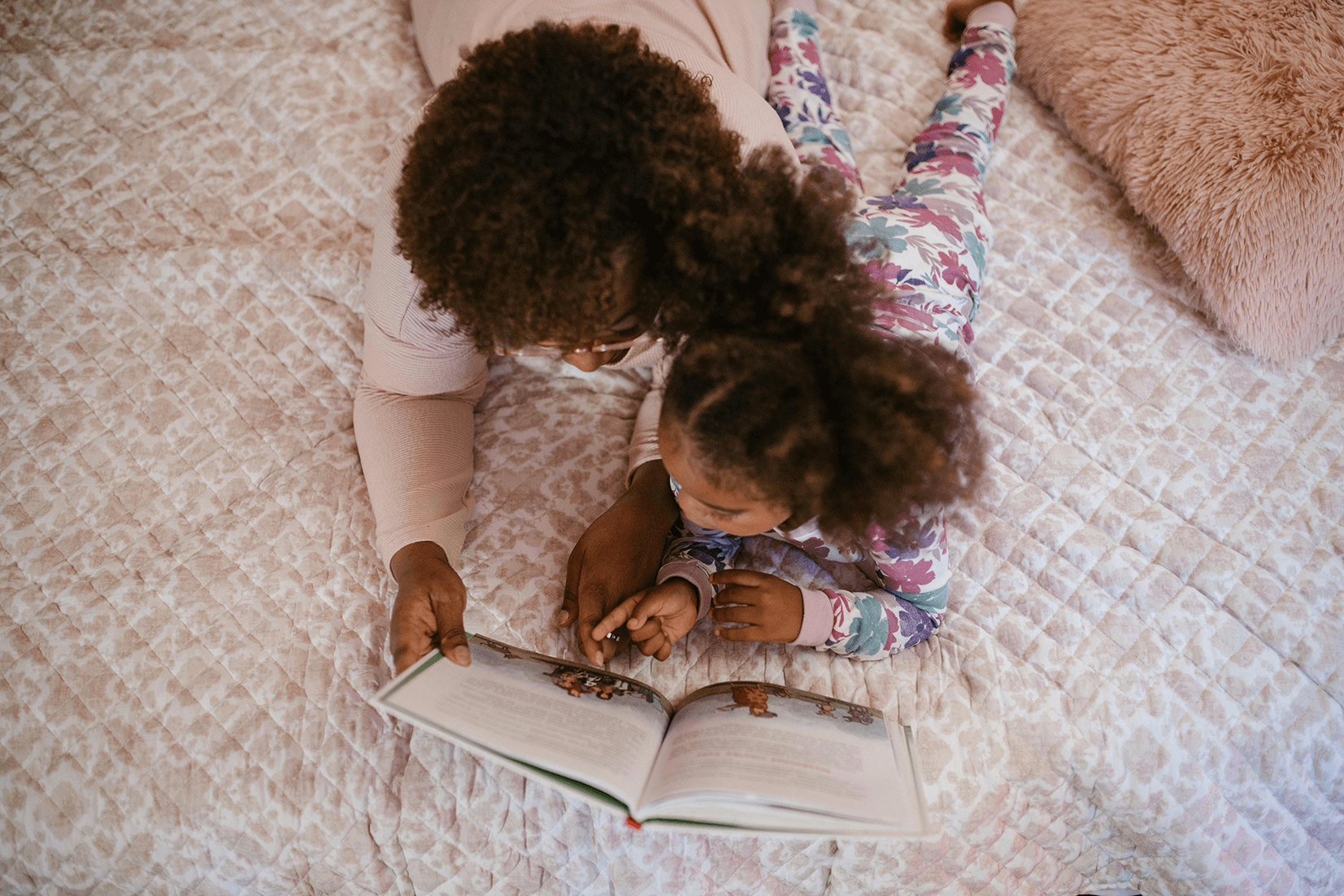 A mom and daughter reading a book together on a bed.